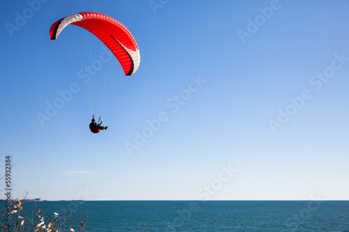 Photo: Red paraglider flying in blue sky over the ocean. Red paraglider flying in blue sky over the ocean.