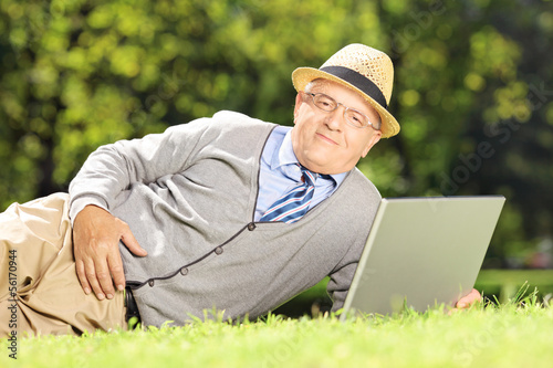 Photo: Senior man with hat lying on a grass working on a laptop in park Senior man with hat lying on a grass working on a laptop in park
