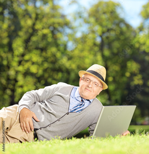 Photo: Senior man with hat lying on a green grass and working on a lapt Senior man with hat lying on a green grass and working on a lapt