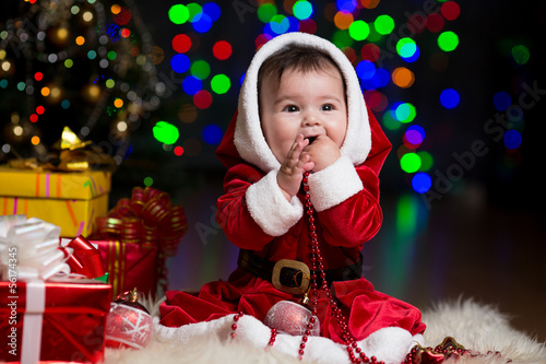 Photo: kid girl dressed as Santa Claus near Christmas tree with gifts kid girl dressed as Santa Claus near Christmas tree with gifts