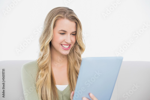 Photo: Smiling pretty blonde using her tablet sitting on cosy sofa Smiling pretty blonde using her tablet sitting on cosy sofa