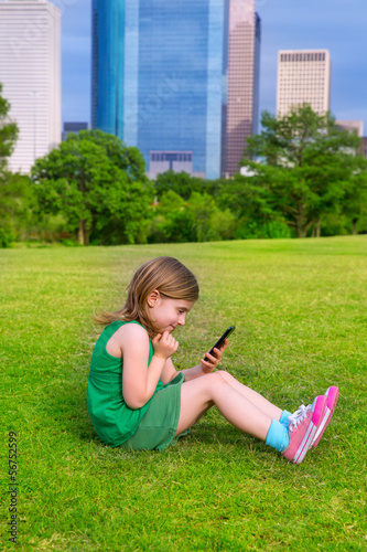 Photo: Blond kid girl playing with smartphone sitting on park lawn at c Blond kid girl playing with smartphone sitting on park lawn at c