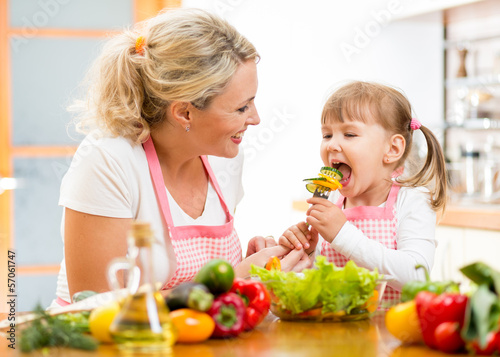 Photo: mother feeding kid daughter vegetables in kitchen mother feeding kid daughter vegetables in kitchen
