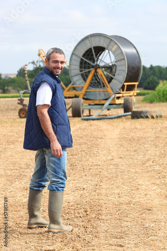 Photo: Famer stood in field machinery in background Famer stood in field machinery in background