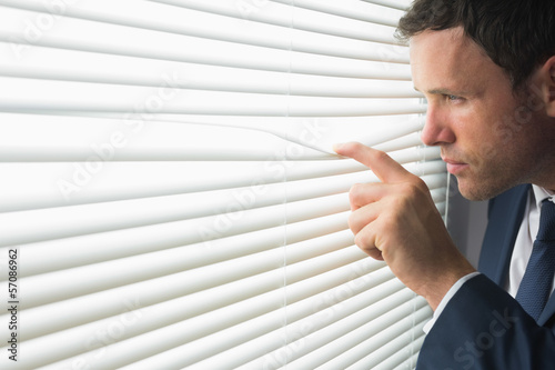 Photo: Serious handsome businessman looking through roller blind Serious handsome businessman looking through roller blind