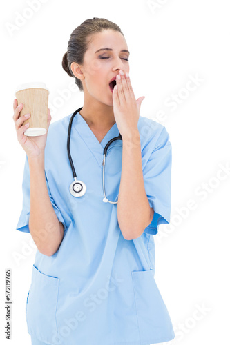 Photo: Tired brown haired nurse in blue scrubs yawning and holding a cu Tired brown haired nurse in blue scrubs yawning and holding a cu