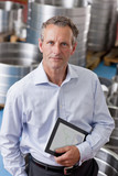 Photo: Portrait of confident businessman holding digital tablet among steel roller bearings in manufacturing plant Portrait of confident businessman holding digital tablet among steel roller bearings in manufacturing plant