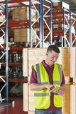 Photo: Smiling worker in reflector-vest using digital tablet in front of crates in warehouse Smiling worker in reflector-vest using digital tablet in front of crates in warehouse