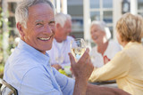 Photo: Portrait of smiling senior man drinking white wine on patio with friends in background Portrait of smiling senior man drinking white wine on patio with friends in background