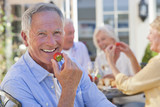 Photo: Portrait of smiling senior man eating strawberry on patio with friends enjoying wine and lunch in background Portrait of smiling senior man eating strawberry on patio with friends enjoying wine and lunch in background