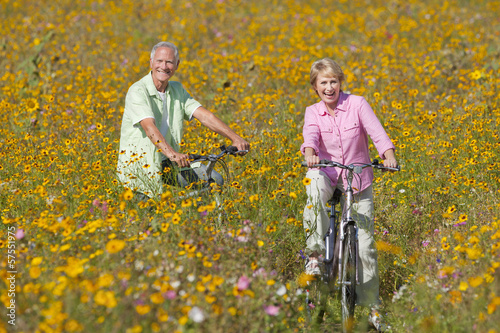 Photo: Happy senior couple riding bicycles in sunny wildflower meadow Happy senior couple riding bicycles in sunny wildflower meadow