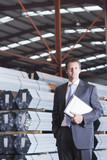 Photo: Portrait of confident businessman with paperwork in front of steel tubing in warehouse Portrait of confident businessman with paperwork in front of steel tubing in warehouse
