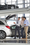 Photo: Salesman and couple with brochure looking into hatchback of car in car dealership showroom Salesman and couple with brochure looking into hatchback of car in car dealership showroom