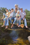 Photo: Portrait of smiling multi-generation family sitting barefoot on footbridge over stream Portrait of smiling multi-generation family sitting barefoot on footbridge over stream