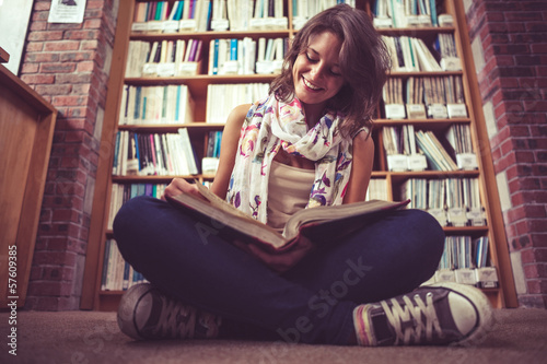 Photo: Happy female student against bookshelf reading a book on the lib Happy female student against bookshelf reading a book on the lib