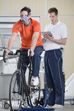 Photo: Sports scientist with digital tablet monitoring cyclist with mask on exercise bike in laboratory Sports scientist with digital tablet monitoring cyclist with mask on exercise bike in laboratory