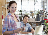 Photo: Portrait of smiling woman holding digital tablet with man listening to headphones and using laptop in background Portrait of smiling woman holding digital tablet with man listening to headphones and using laptop in background