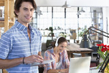 Photo: Portrait of smiling man holding digital tablet with woman listening to headphones and using laptop in background Portrait of smiling man holding digital tablet with woman listening to headphones and using laptop in background