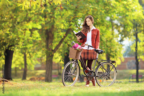 Photo: Young female with bicycle in a park reading book Young female with bicycle in a park reading book