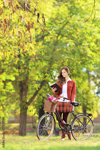Photo: Young woman with bicycle in a park reading book Young woman with bicycle in a park reading book
