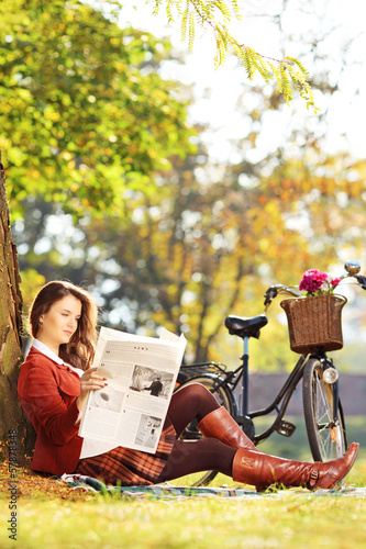 Photo: Young woman with bike sitting and reading a newspaper in park Young woman with bike sitting and reading a newspaper in park