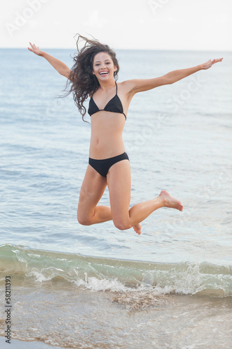 Photo: Cheerful young woman in bikini jumping at beach Cheerful young woman in bikini jumping at beach