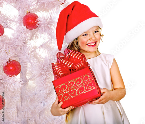 Photo: Child in Santa hat with gift box near white Christmas tree. Child in Santa hat with gift box near white Christmas tree.