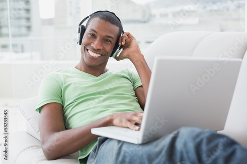 Photo: Smiling young Afro man with headphones using laptop on sofa Smiling young Afro man with headphones using laptop on sofa