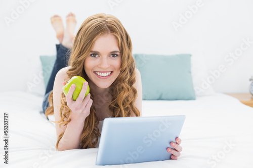 Photo: Casual blond using tablet PC while holding an apple in bed Casual blond using tablet PC while holding an apple in bed