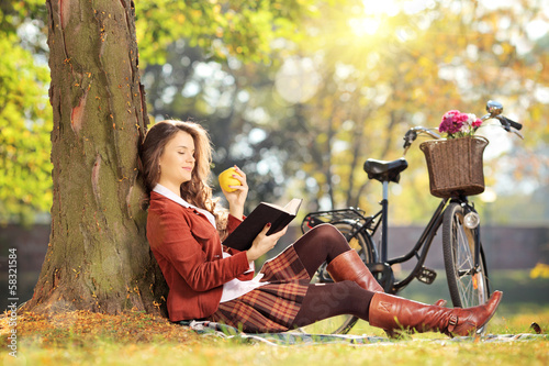 Photo: Young female in park reading a book and eating on a sunny day Young female in park reading a book and eating on a sunny day