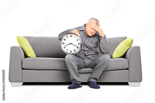 Photo: Mature man in pajamas holding a clock and having a headache Mature man in pajamas holding a clock and having a headache