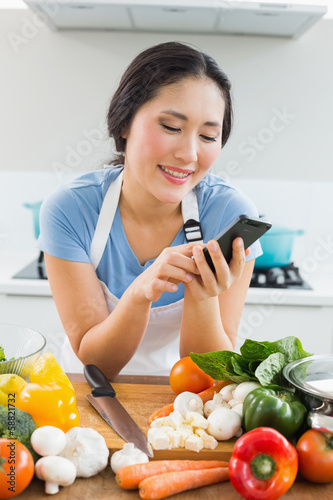 Photo: Woman text messaging in front of vegetables in kitchen Woman text messaging in front of vegetables in kitchen