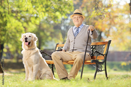 Photo: Senior man seated on a bench with a dog relaxing in a park Senior man seated on a bench with a dog relaxing in a park