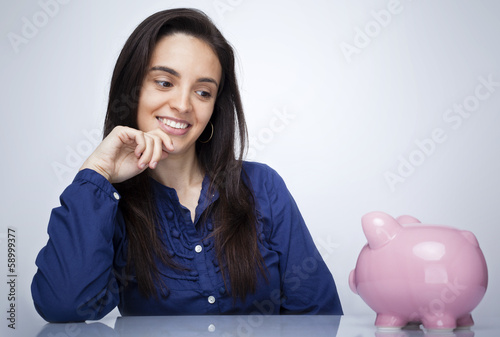 Photo: Happy smiling woman looking to a piggy bank against gray backgro Happy smiling woman looking to a piggy bank against gray backgro