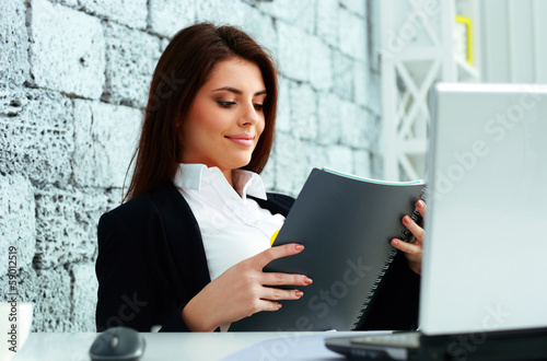 Photo: Young beautiful businesswoman reading the notebook in office Young beautiful businesswoman reading the notebook in office
