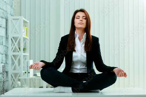 Photo: Young beautiful businesswoman meditates on the table in office Young beautiful businesswoman meditates on the table in office