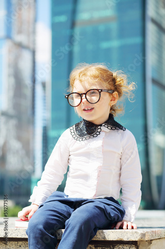 Photo: Little cute girl in glasses sits on border near skyskraper Little cute girl in glasses sits on border near skyskraper