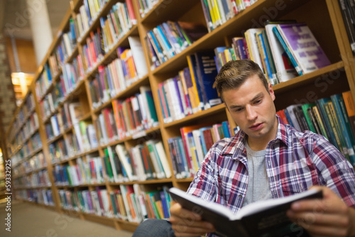 Photo: Focused young student sitting on library floor reading book Focused young student sitting on library floor reading book