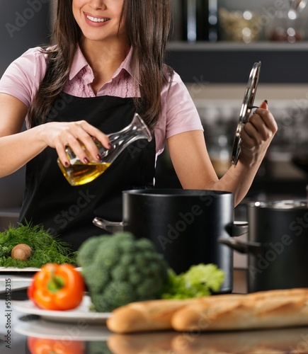 Photo: Smiling young woman adding oil to pot on a modern kitchen Smiling young woman adding oil to pot on a modern kitchen