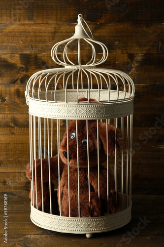 Photo: Toy bear in decorative cage on wooden background Toy bear in decorative cage on wooden background