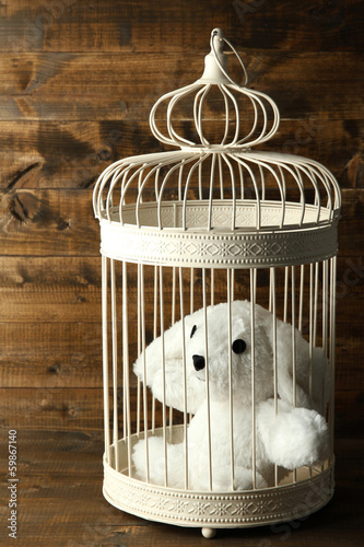 Photo: Toy rabbit in decorative cage on wooden background Toy rabbit in decorative cage on wooden background