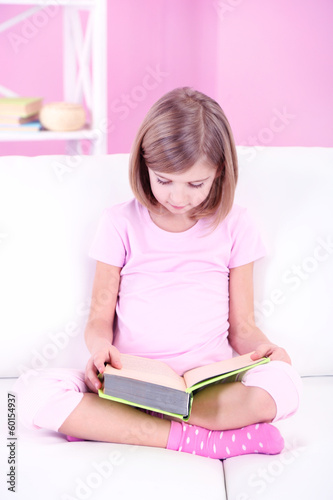 Photo: Little girl reading book sitting on sofa in room Little girl reading book sitting on sofa in room
