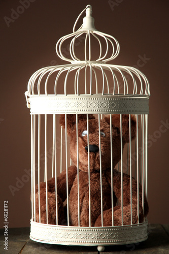 Photo: Toy bear in decorative cage on wooden table, on brown Toy bear in decorative cage on wooden table, on brown