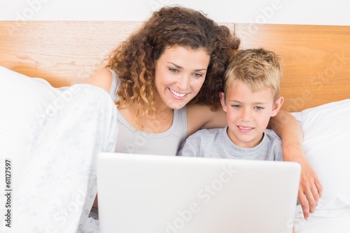 Photo: Cheerful mother and son sitting on bed using laptop Cheerful mother and son sitting on bed using laptop