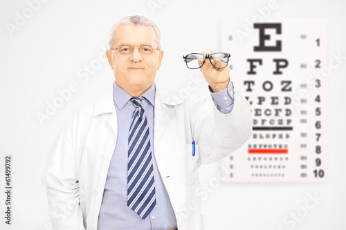 Photo: Male optician holding glasses in front of an eye chart Male optician holding glasses in front of an eye chart