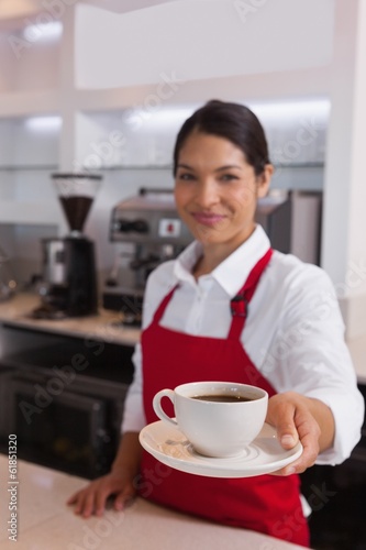 Photo: Pretty barista offering cup of coffee smiling at camera Pretty barista offering cup of coffee smiling at camera