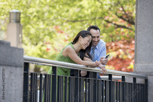 Foto: A Couple, Man And Woman Checking A Smart Phone, Under The Shade Of Trees In The Park. A Couple, Man And Woman Checking A Smart Phone, Under The Shade Of Trees In The Park.