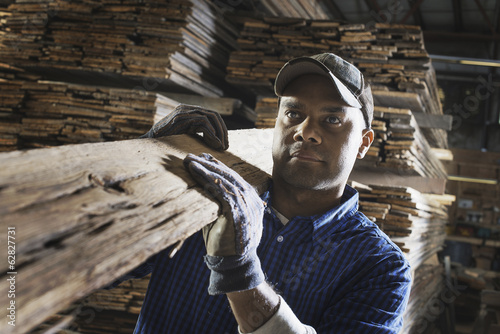 Foto: A heap of recycled reclaimed timber planks of wood. Environmentally responsible reclamation in a timber yard. A man carrying a large plank of splintered rough wood. A heap of recycled reclaimed timber planks of wood. Environmentally responsible reclamation in a timber yard. A man carrying a large plank of splintered rough wood.