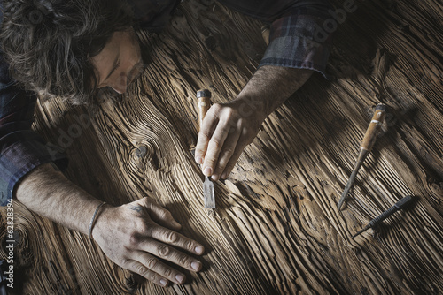 Foto: A man working in a reclaimed lumber yard workshop. Holding tools and sanding knotted and uneven piece of wood. A man working in a reclaimed lumber yard workshop. Holding tools and sanding knotted and uneven piece of wood.