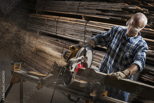 Foto: A reclaimed lumber workshop. A man in protective eye goggles using a circular saw to cut timber. A reclaimed lumber workshop. A man in protective eye goggles using a circular saw to cut timber.